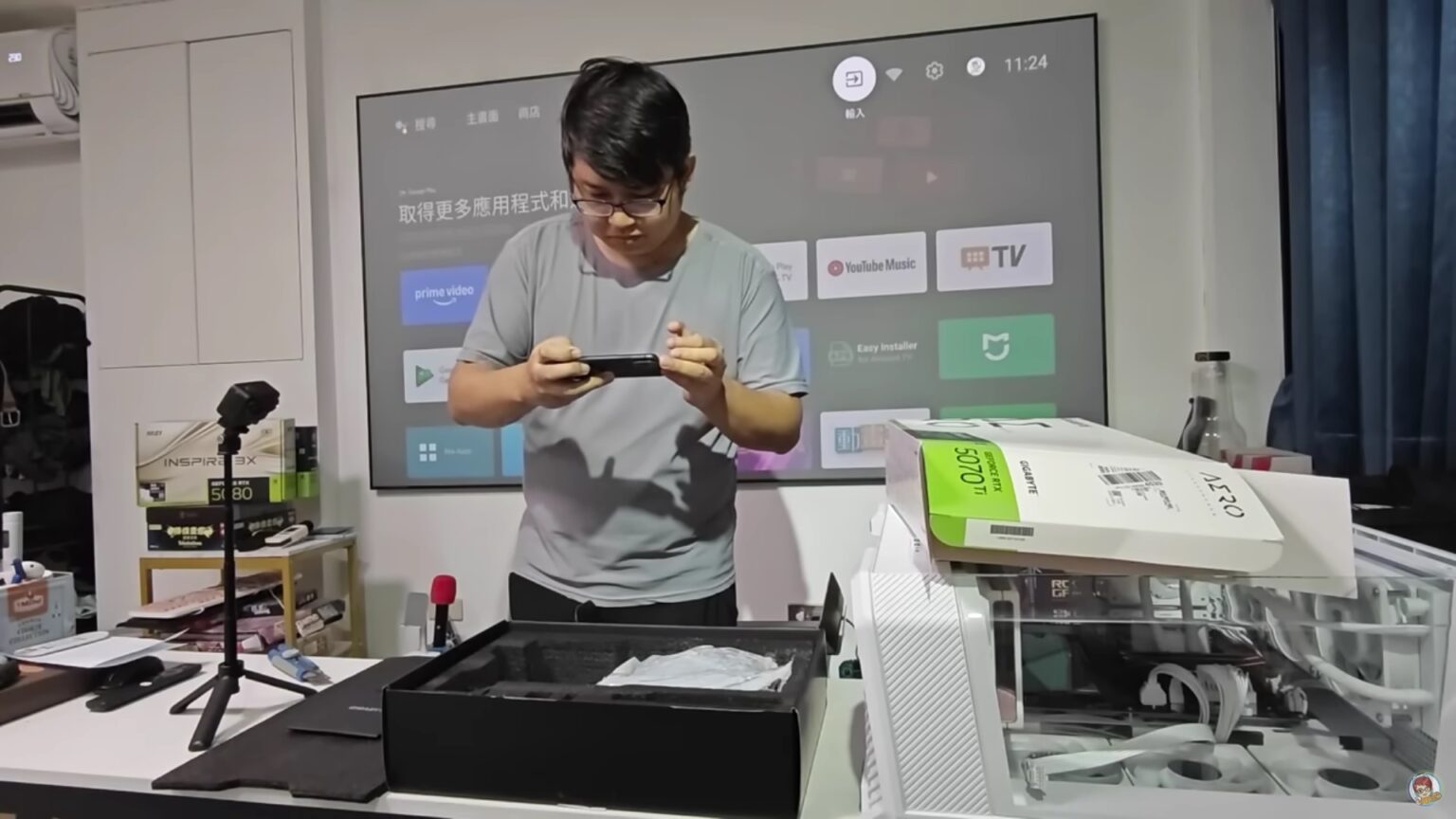 Homem de óculos segura um dispositivo preto em frente a uma TV com Android TV, ao lado de caixa aberta e uma caixa de placa de vídeo sobre uma mesa