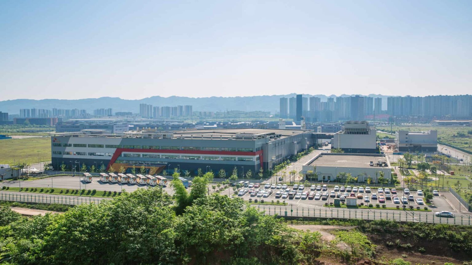 Vista aérea de grande complexo industrial com galpão de fachada colorida, caminhões estacionados, pátio com carros e prédios residenciais ao fundo