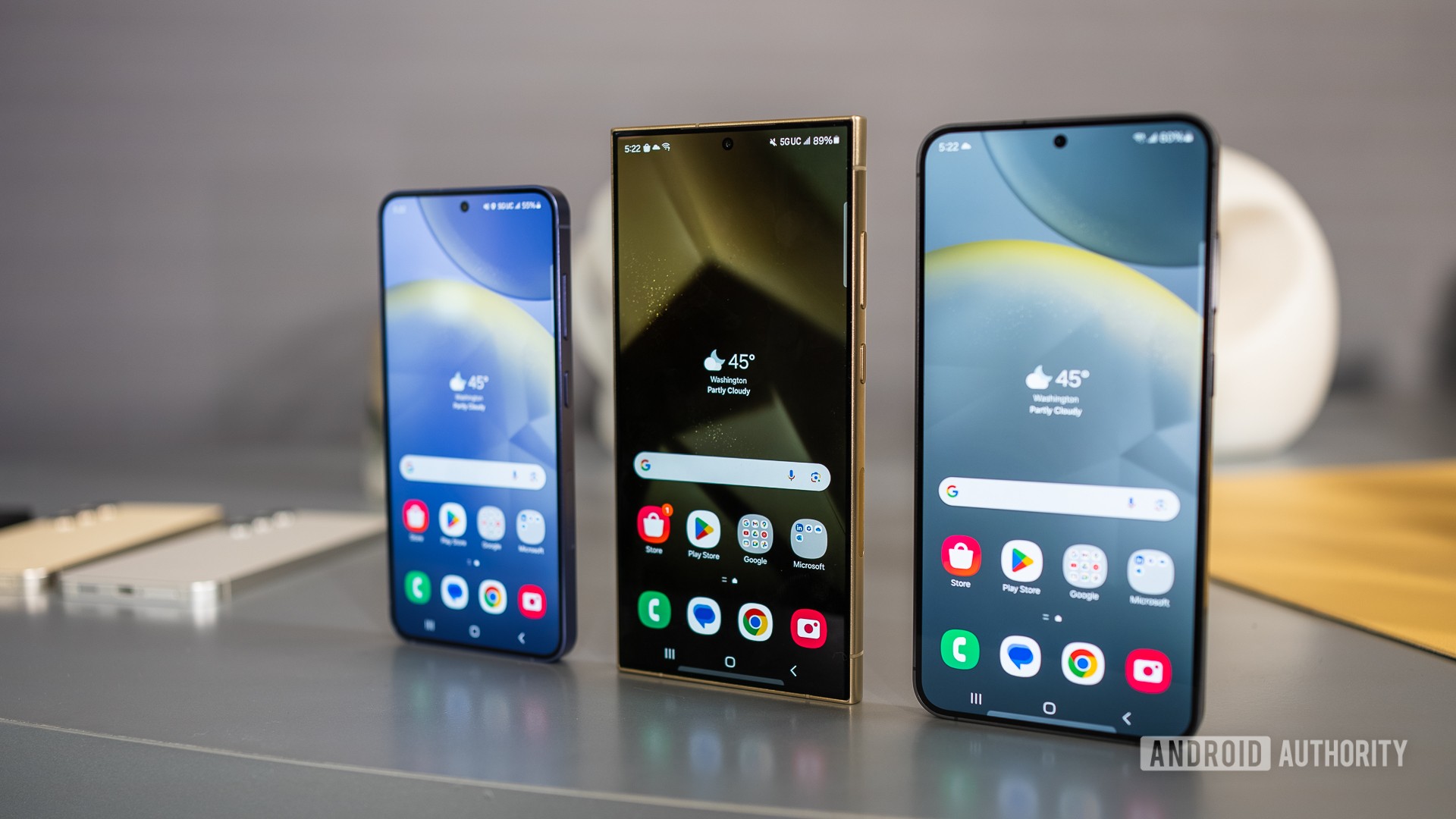 Samsung começa a liberar a One UI 8 para o Galaxy S24 1 Três smartphones Samsung Galaxy S24 alinhados sobre superfície cinza com telas iniciais exibindo widget de clima e ícones
