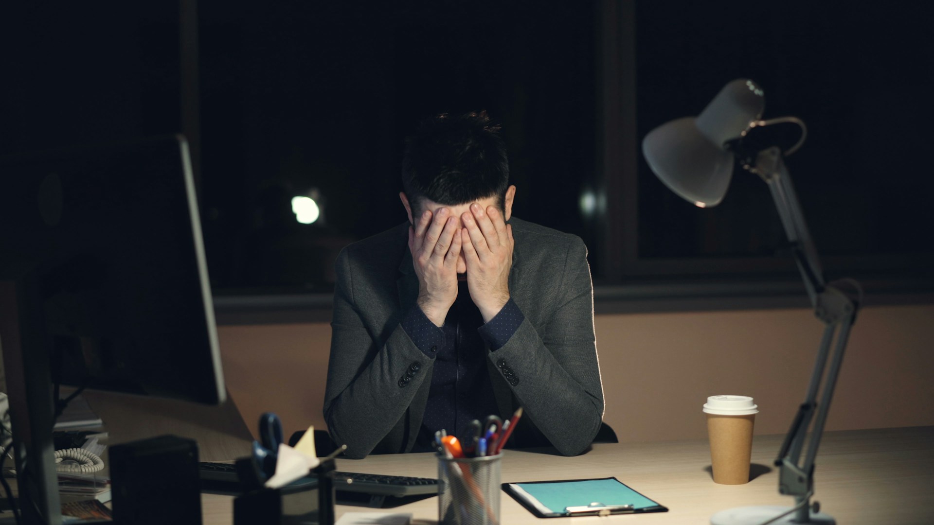 Workslop: como a IA preguiçosa está sobrecarregando empresas e colegas 1 Homem cansado segurando o rosto com as mãos em escritório iluminado à noite com itens de trabalho sobre a mesa