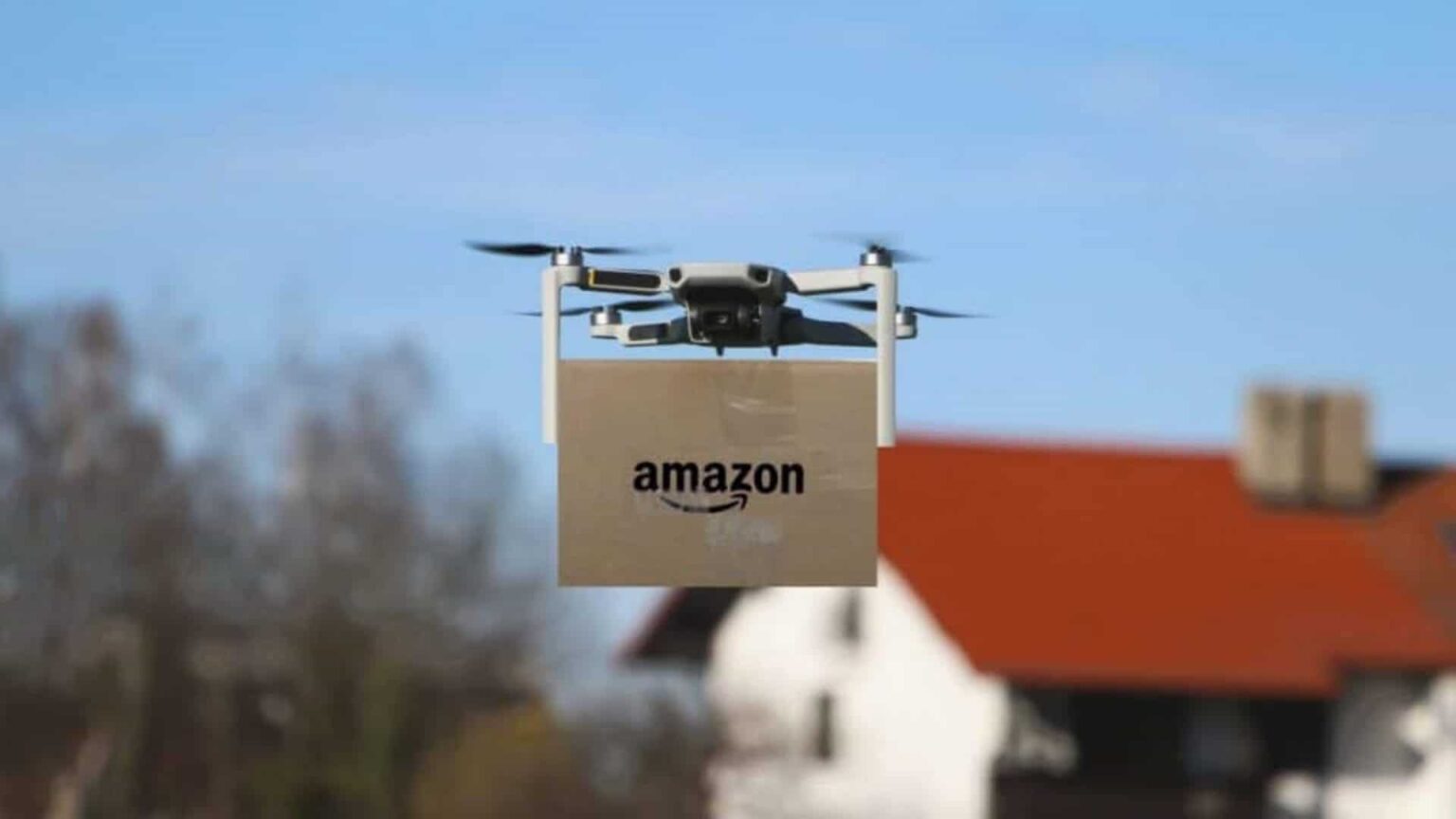 Drone cinza carregando caixa de papelão com logo da Amazon sobre casa com telhado vermelho ao fundo