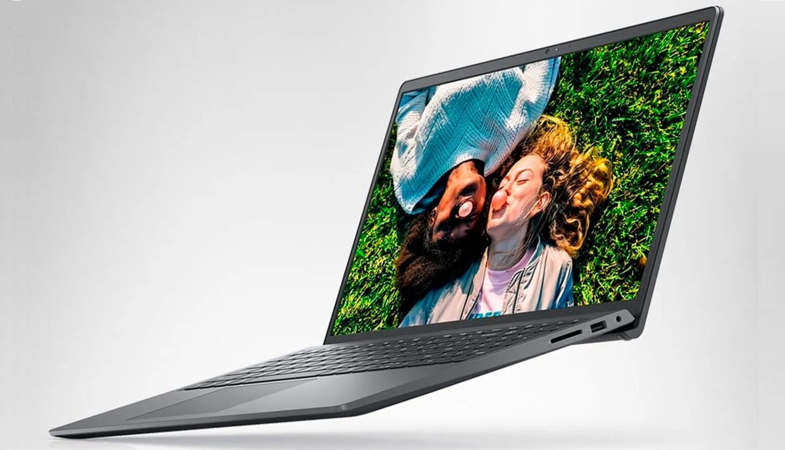 Notebook Dell Inspiron cinza em ângulo, com tela exibindo um casal deitado na grama sorrindo, sobre fundo claro