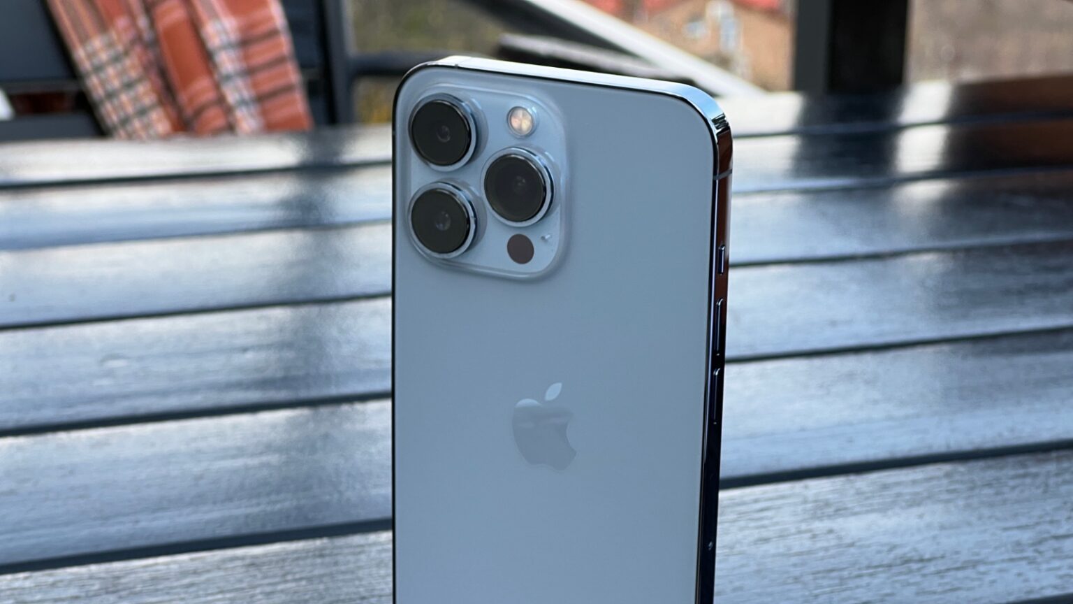 iPhone 13 Pro azul-sierra visto pela traseira, com o conjunto triplo de câmeras e o logo da Apple, apoiado sobre uma mesa de madeira ao ar livre