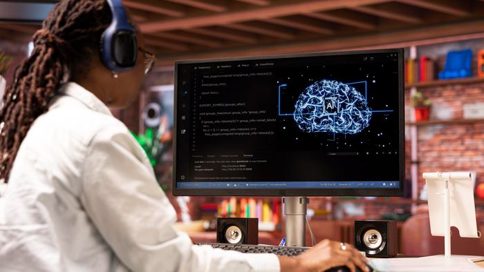 Ex-pesquisadores da OpenAI, Google e Meta lançam a Periodic Labs para transformar a ciência com IA 2 Pessoa sentada em espaço de trabalho tecnológico com fones de ouvido azuis e monitor mostrando código e cérebro em neon azul