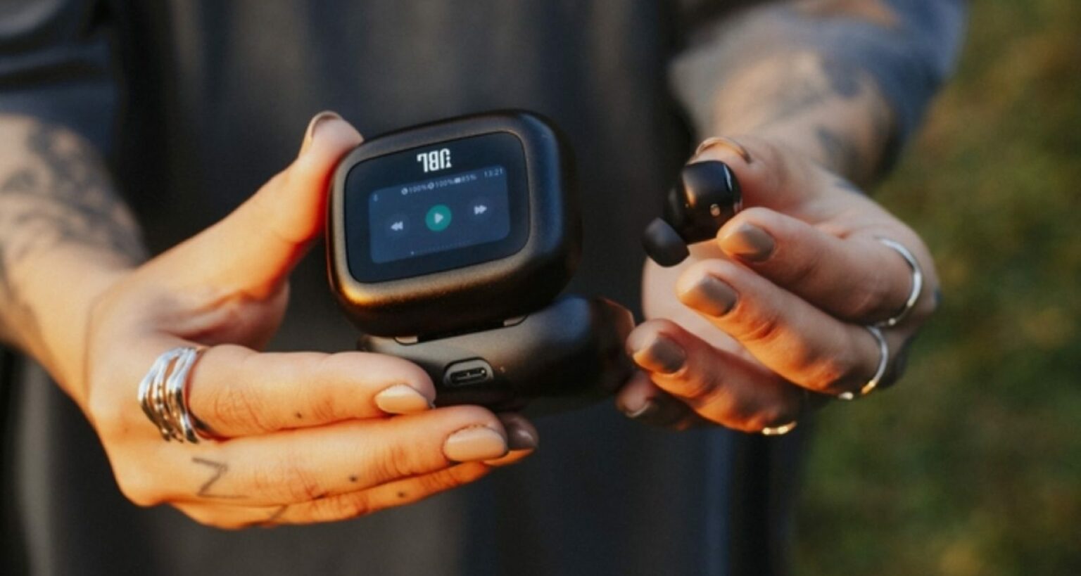 Pessoa segurando case de fone de ouvido JBL preto com tela touchscreen mostrando controles de reprodução, e um earbud ao lado