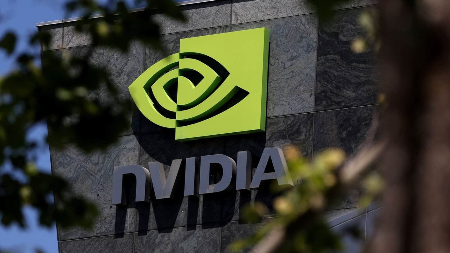 Logotipo da Nvidia em letras cinzas e símbolo verde na fachada de um prédio de pedra escura, com folhagem desfocada em primeiro plano