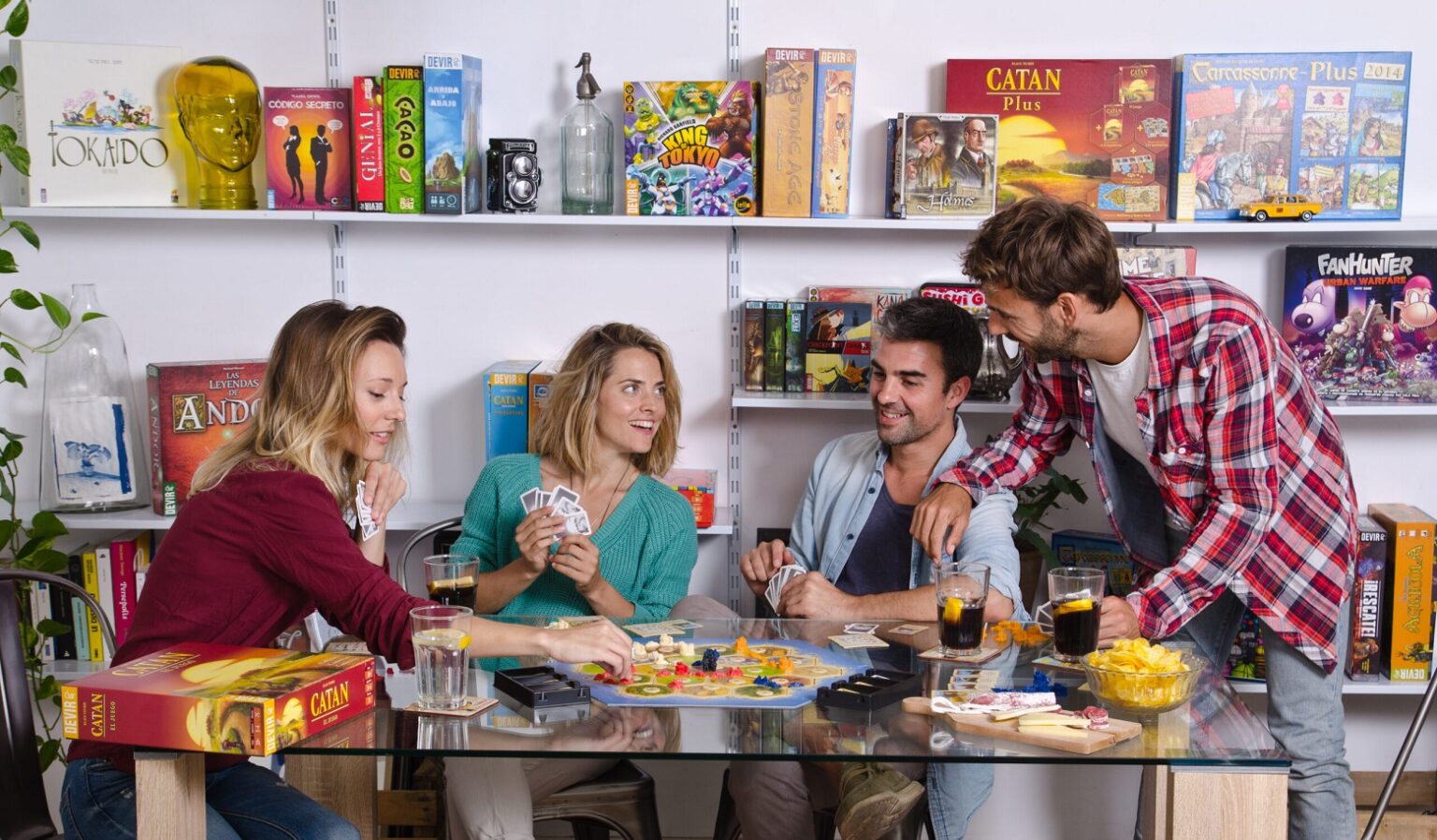 Quatro amigos jogam tabuleiro em uma mesa de vidro, com caixas de Catan e outros jogos expostas nas prateleiras ao fundo