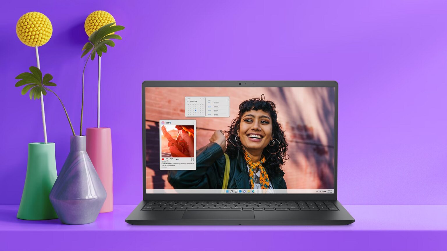 Notebook Dell Inspiron 15 preto sobre bancada roxa, com tela exibindo uma mulher sorrindo ao lado de widgets de calendário e rede social