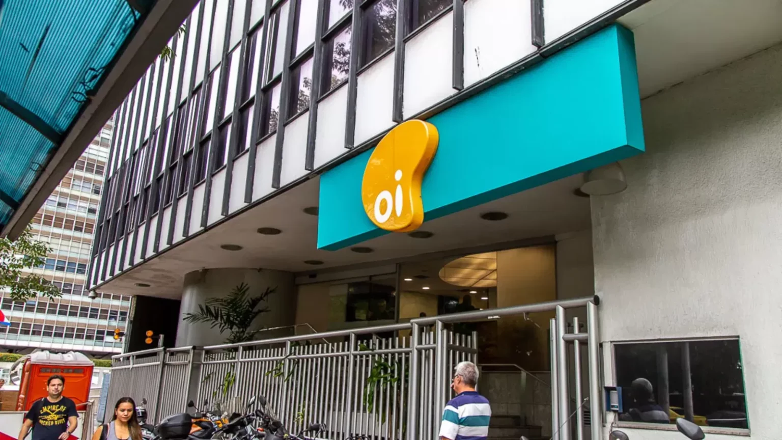 Fachada de prédio da operadora Oi, com placa turquesa e logotipo amarelo, em uma rua movimentada com pedestres e motos estacionadas