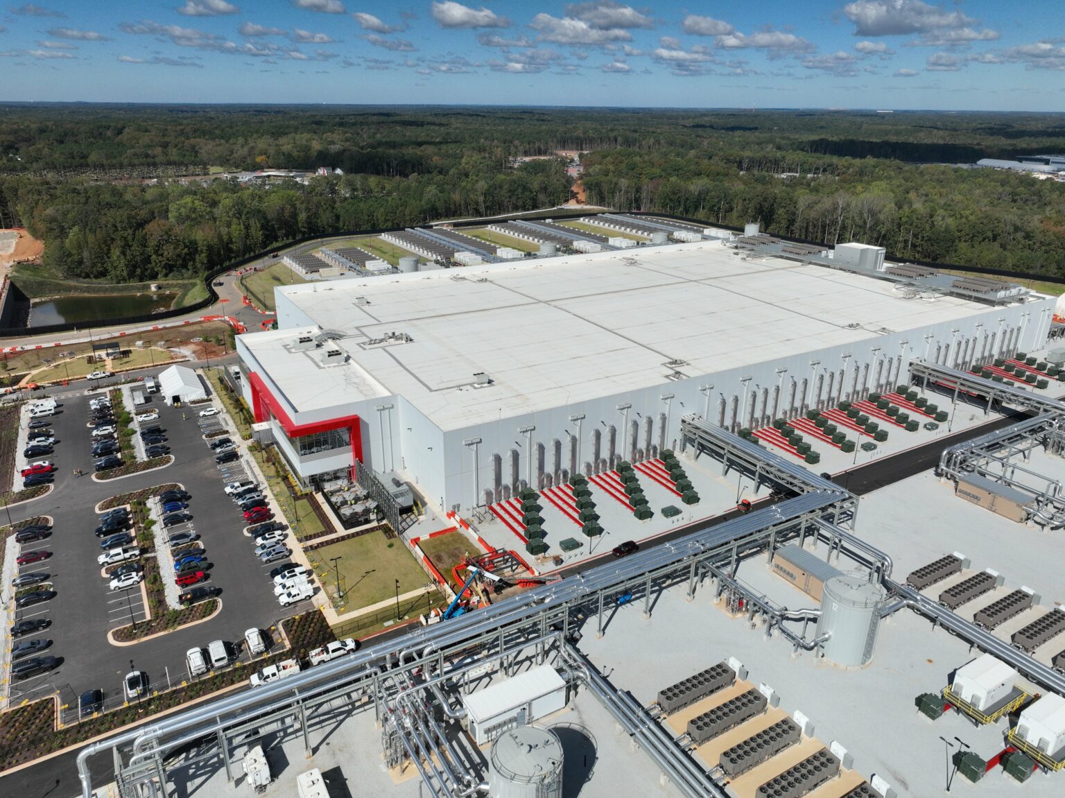 Vista aérea de um grande datacenter branco com faixas vermelhas, estacionamento à esquerda e equipamentos de refrigeração instalados no pátio externo, cercado…