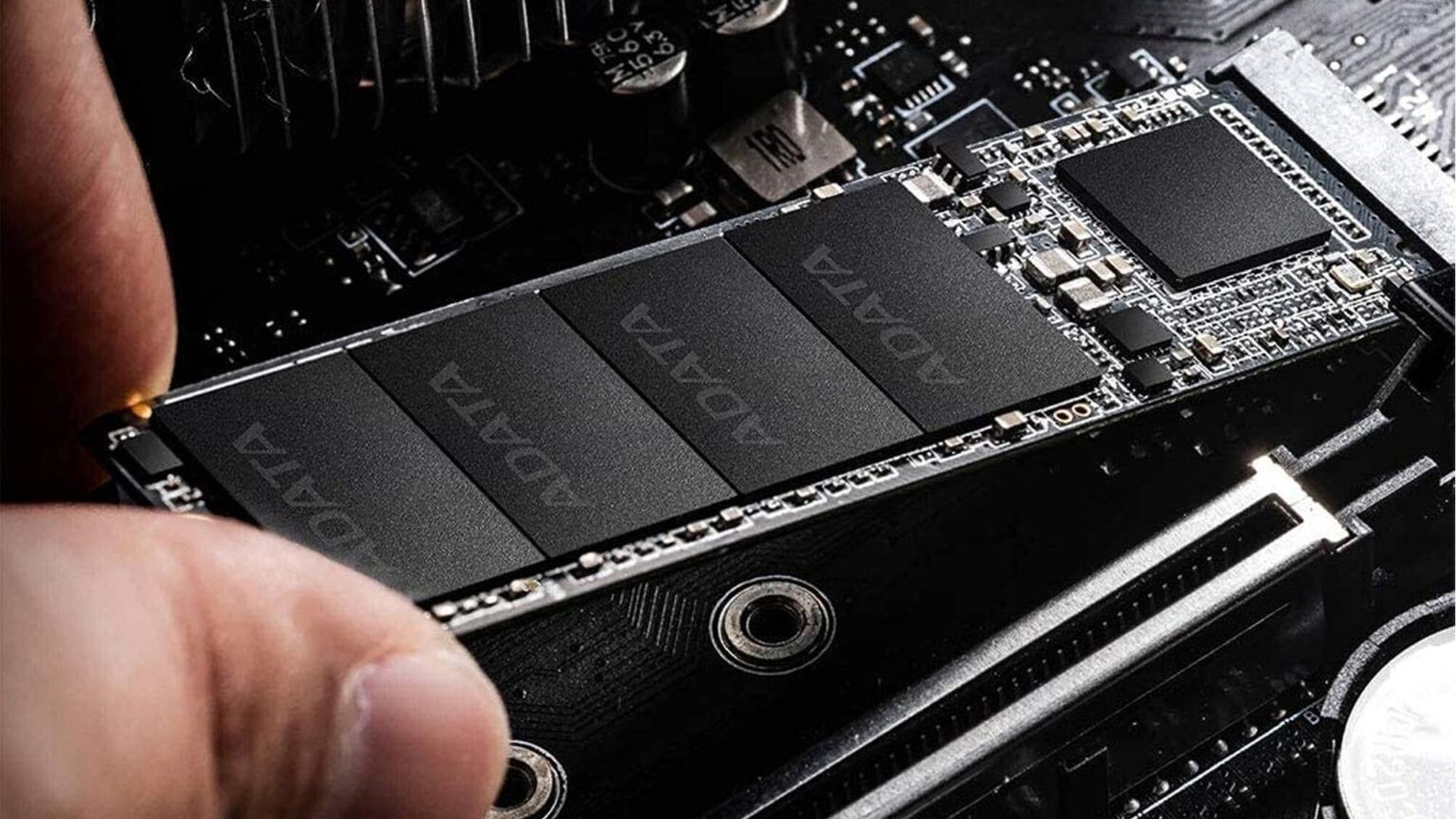 Mão segurando um SSD M.2 preto da Adata instalado em placa-mãe, com o nome da marca visível sobre os dissipadores