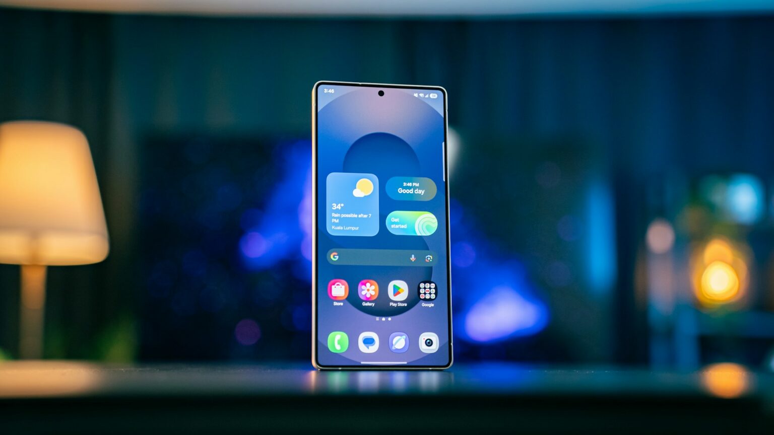 Smartphone Samsung em pé sobre uma mesa, exibindo a tela inicial com widgets de clima e Google, sob iluminação ambiente azulada e um abajur desfocado ao fundo