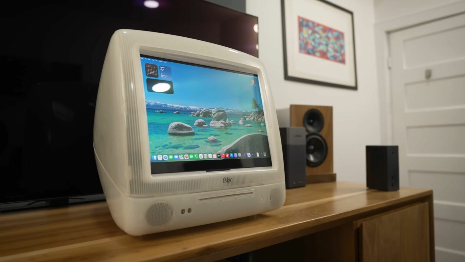iMac G3 bege com tela exibindo área de trabalho de macOS com paisagem de lago, sobre uma mesa de madeira ao lado de caixas de som