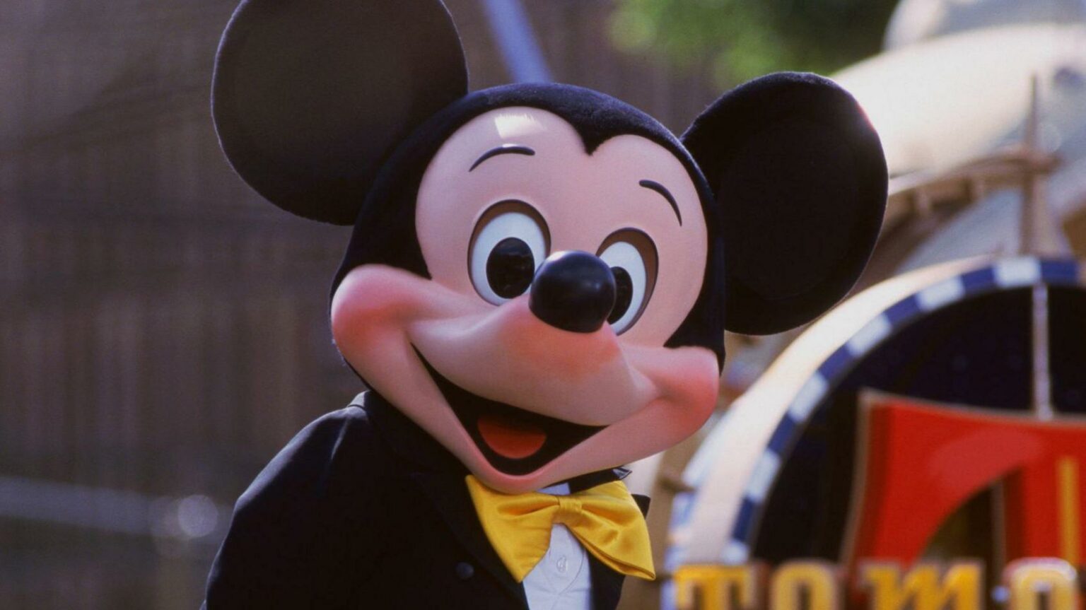 Mascote com cabeça grande, orelhas redondas pretas, luvas brancas e gravata-borboleta amarela, sorrindo em frente a um trem colorido de parque temático
