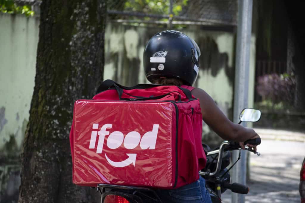 Governo lança grupo para propor regras trabalhistas para entregadores por aplicativo no Brasil 1 Entregadora de moto com mochila térmica vermelha do iFood em cenário urbano diurno