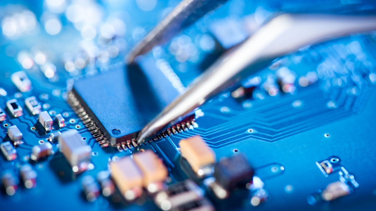 Nova tarifa dos EUA sobre semicondutores chineses entra em vigor só em 2027, mas o impacto já preocupa 1 Close-up macro de microchip preto retangular sobre placa de circuito azul com pinça metálica tocando pinos
