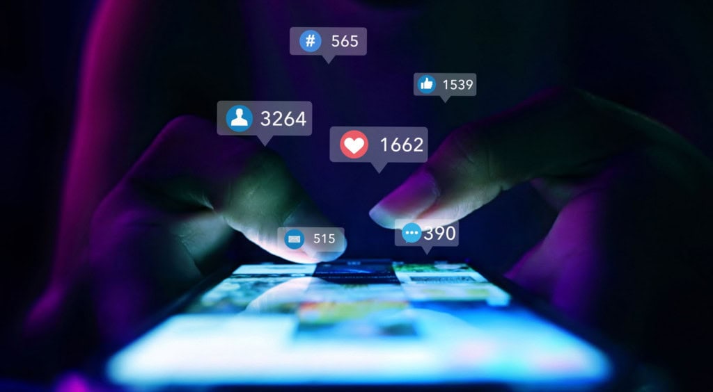 Nova regra dos EUA pode exigir histórico das redes sociais dos últimos 5 anos para para liberar entrada no país 2 Mãos usando smartphone com feed de rede social e ícones de notificações em ambiente noturno roxo e azul