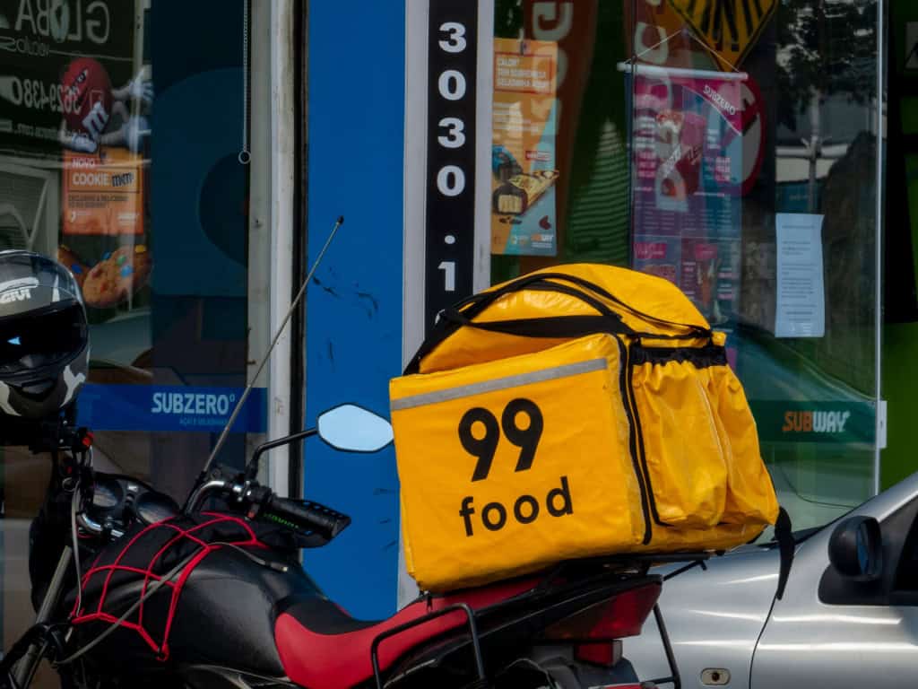 Motocicleta estacionada com mochila térmica amarela da 99Food em frente à loja e vitrine urbana