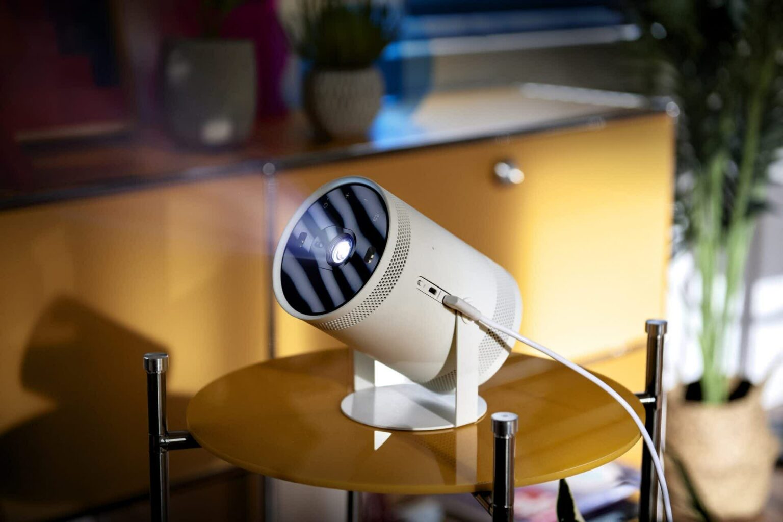 Projetor portátil cilíndrico branco Samsung Freestyle sobre mesa redonda amarela, com cabo USB-C conectado e lente acesa em ambiente de sala