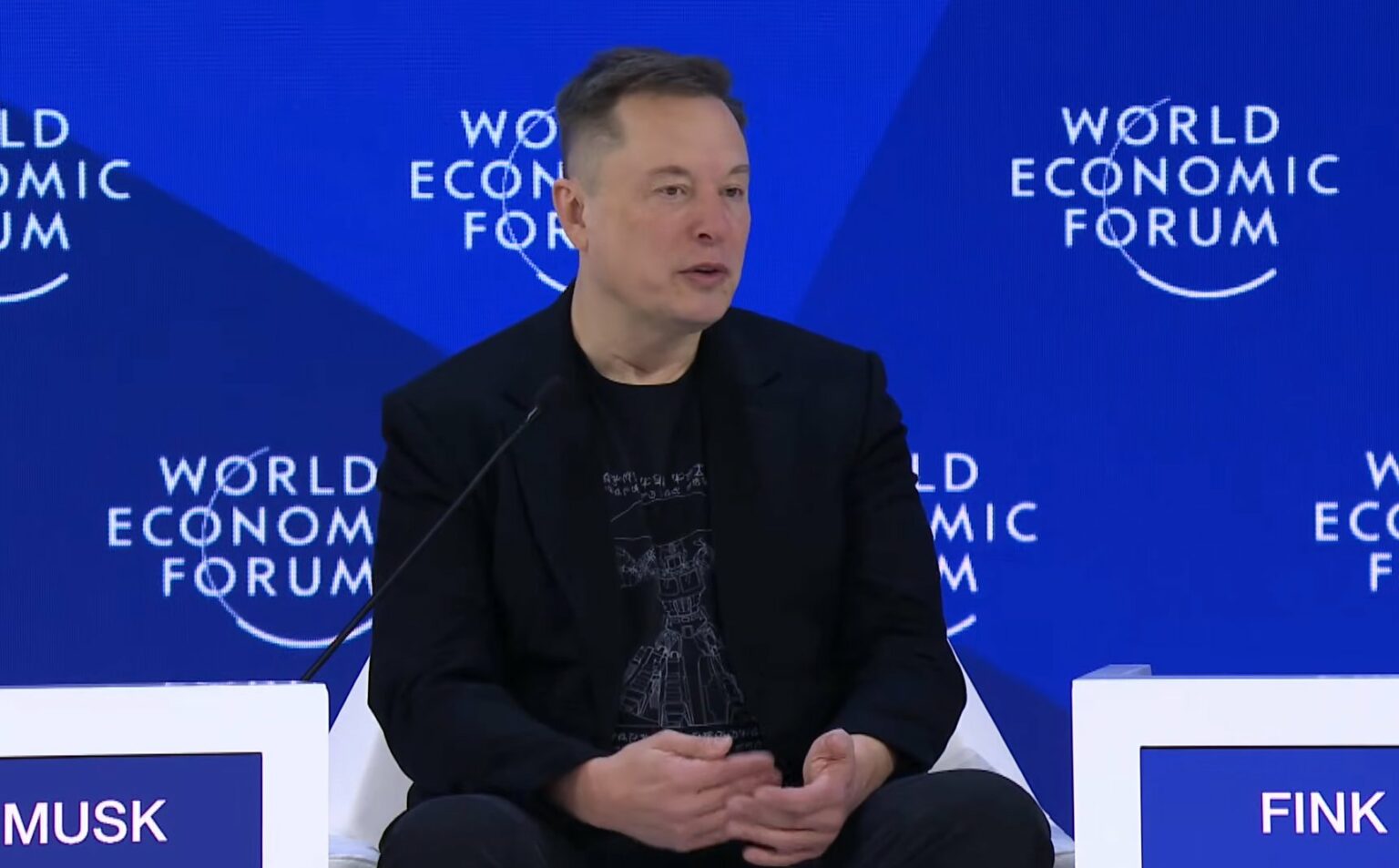 Homem de terno preto sentado em painel do Fórum Econômico Mundial, com placas identificando "MUSK" e "FINK" e o logo do evento ao fundo
