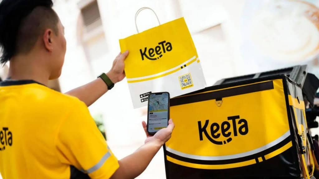 Entregador vestido com uniforme amarelo Keeta segurando sacola de papel com logo Keeta e smartphone com mapa na tela