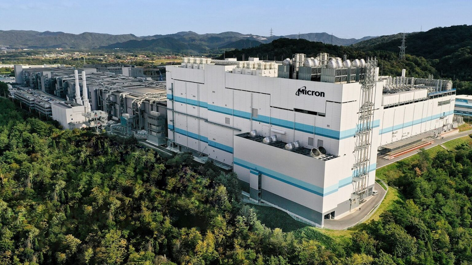 Vista aérea de uma grande fábrica branca com o logotipo Micron no topo, cercada por árvores e montanhas ao fundo