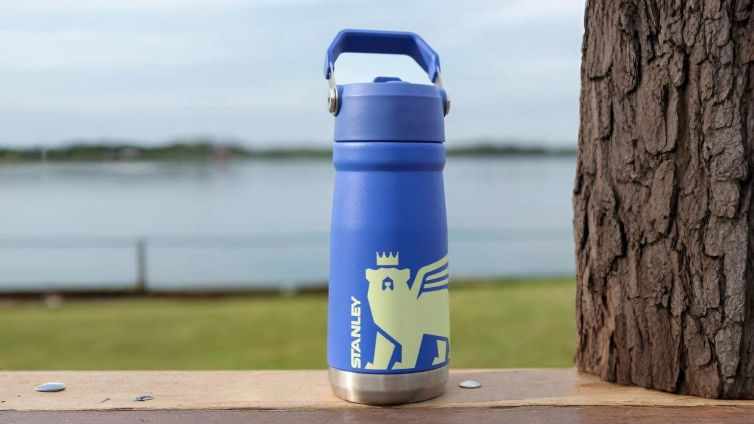 Garrafa térmica azul Stanley com canudo dobrável e alça, estampa de urso com coroa em verde-claro, sobre um banco de madeira com árvore e lago ao fundo