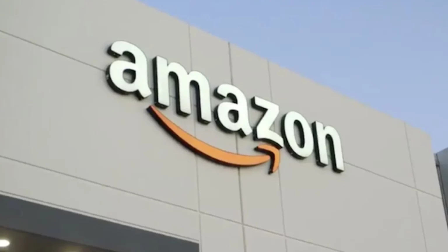 Fachada de prédio cinza com o logotipo da Amazon em letras brancas e a seta laranja em formato de sorriso, sob céu azul