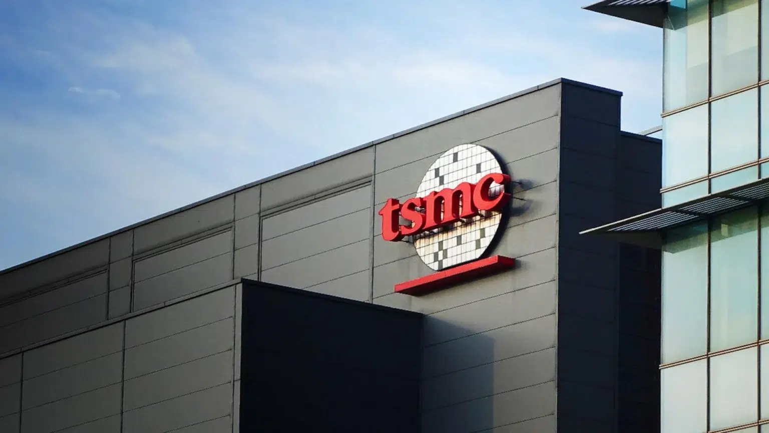 Fachada de prédio cinza com o logotipo vermelho da TSMC, com um disco branco de wafer de silício ao fundo, sob céu azul
