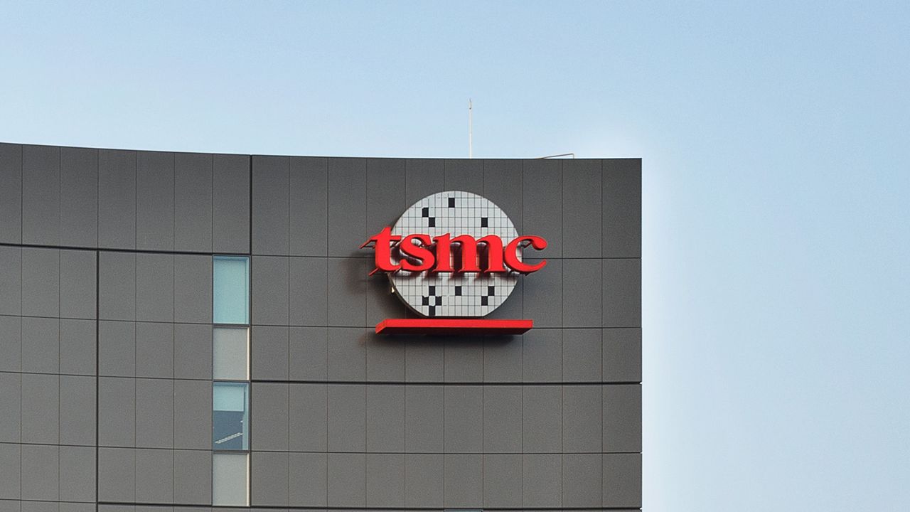 Fachada moderna da TSMC com logotipo vermelho e painéis metálicos cinza sob céu azul claro em dia ensolarado