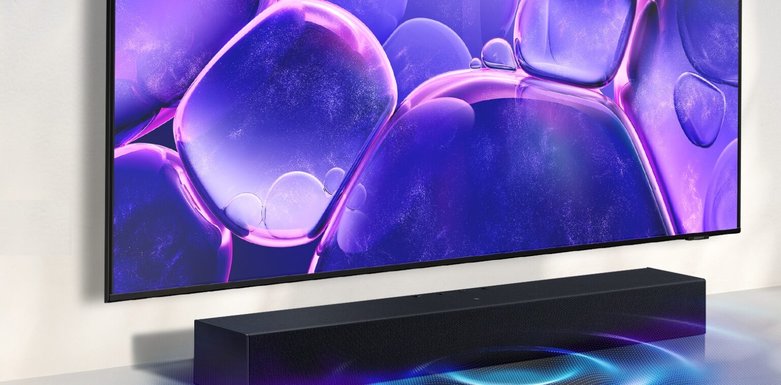 TV Samsung de tela grande exibindo formas roxas e translúcidas, com uma soundbar preta instalada logo abaixo, sobre o rack