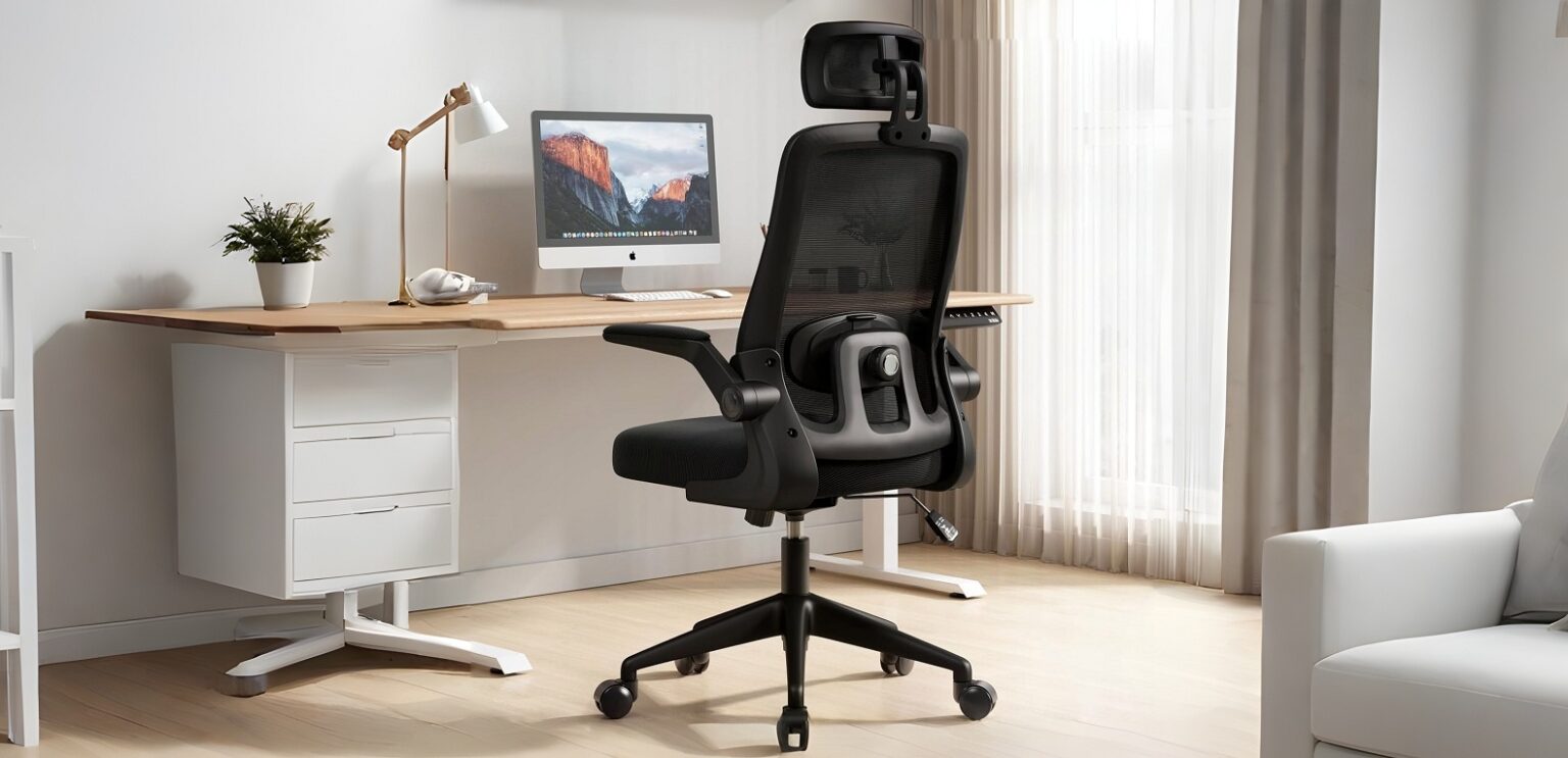 Cadeira de escritório ergonômica preta com encosto em tela mesh, apoio de cabeça e braços articulados, ao lado de uma mesa de madeira com monitor e luminária