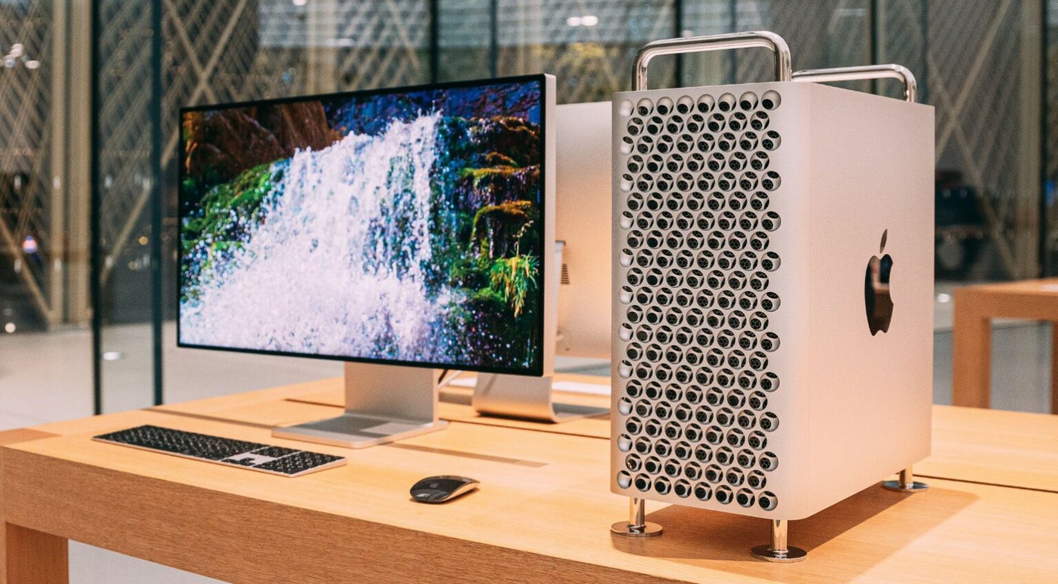 Mac Pro prateado com gabinete perfurado ao lado de monitor Apple Studio Display exibindo cachoeira, com teclado e mouse sobre mesa de madeira em loja Apple