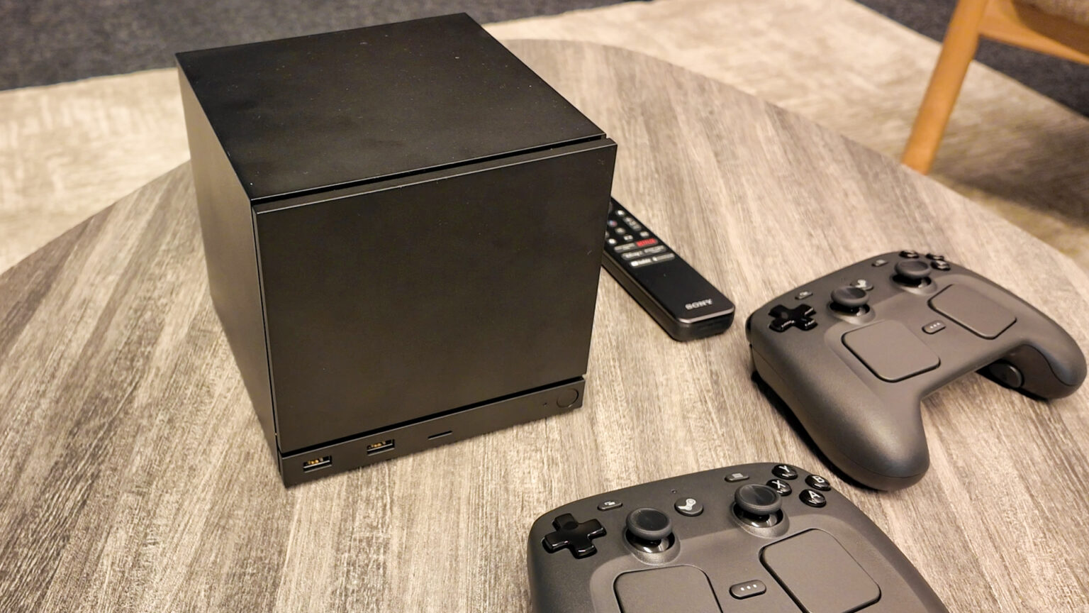Console Steam Machine preto em formato cúbico ao lado de dois controles Steam Controller sobre uma mesa, com um controle remoto Sony ao fundo