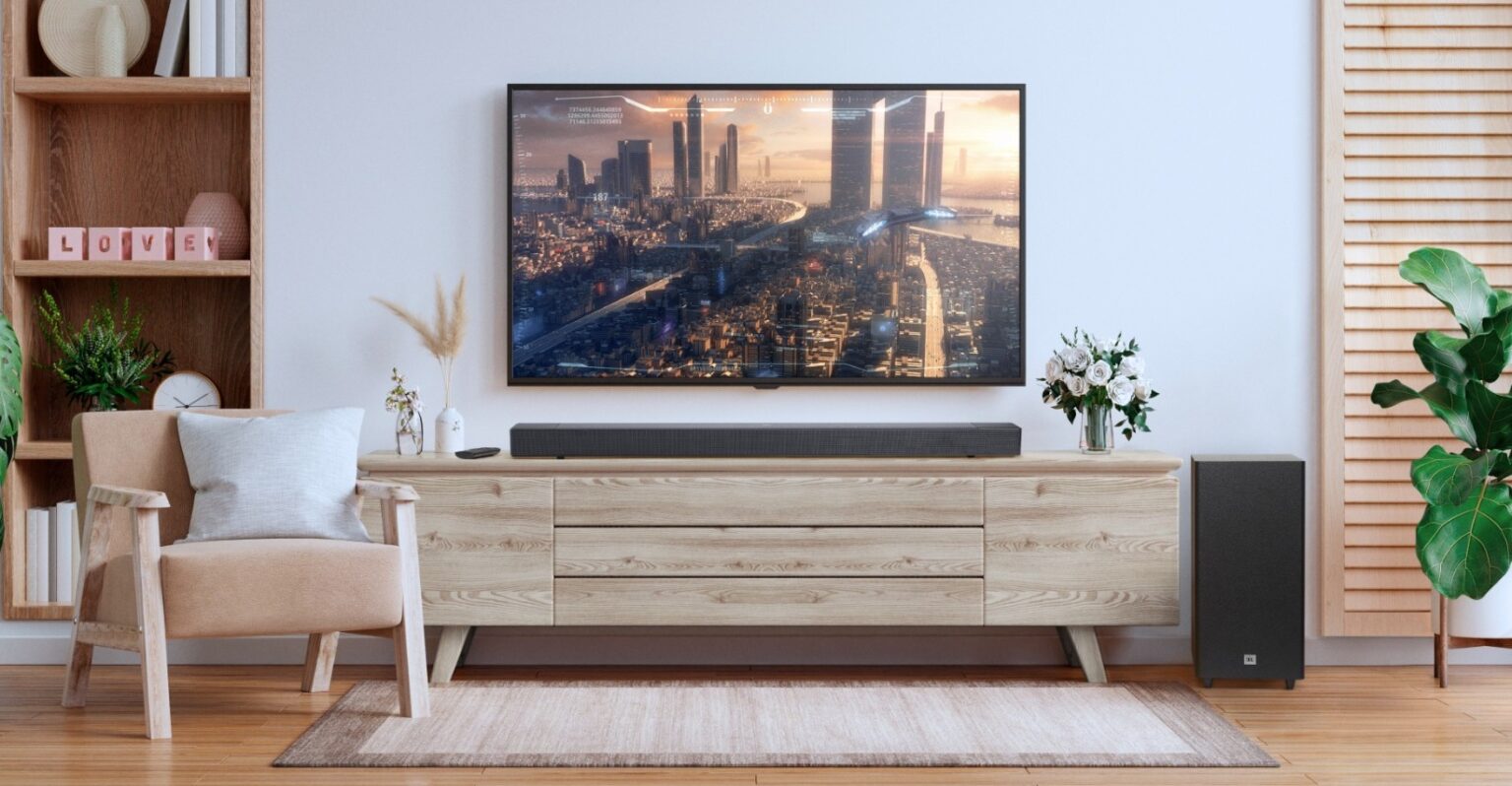 Sala de estar com TV montada na parede, soundbar preta sob ela e subwoofer preto ao lado de um móvel de madeira clara com decoração