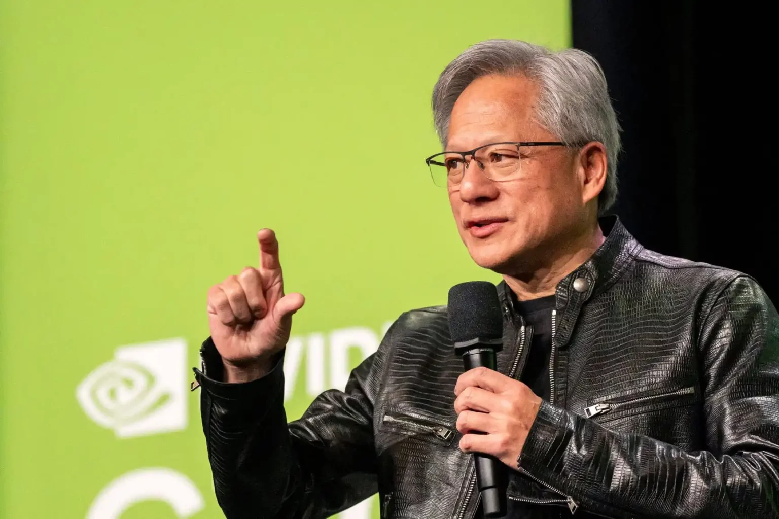 Palestrante de jaqueta de couro preta gesticulando e segurando microfone diante de painel verde com logotipo da NVIDIA