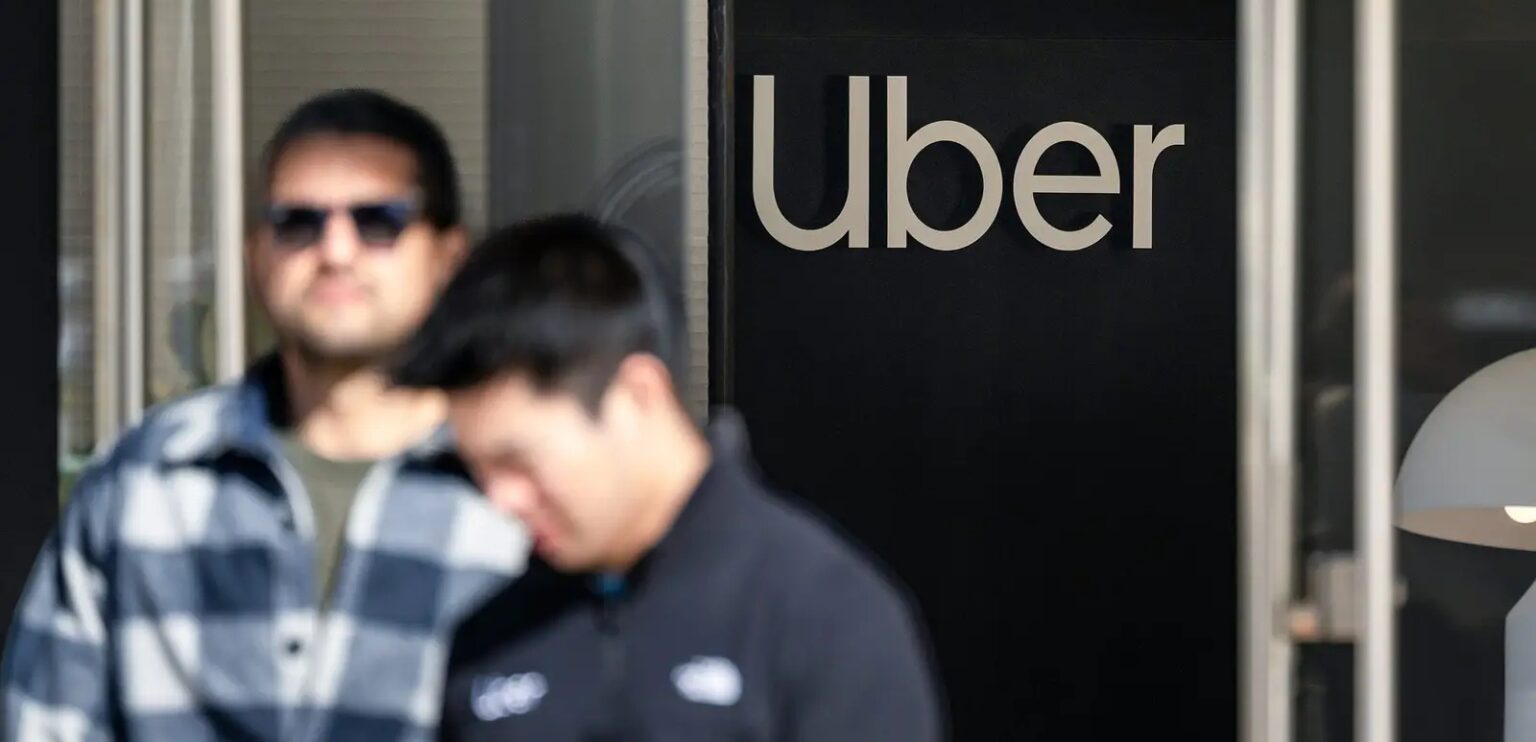 Entrada da sede da Uber com o logotipo na parede escura, em frente a dois homens conversando, um de óculos escuros e camisa xadrez e outro de agasalho escuro