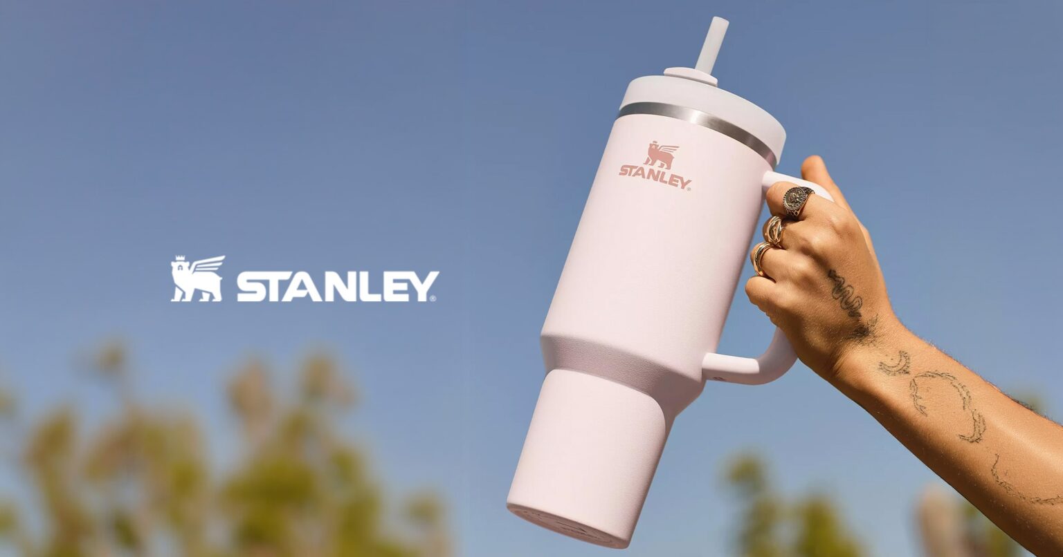Mão segurando garrafa térmica Stanley rosa claro com canudo e alça, em frente a um céu azul; logo Stanley à esquerda