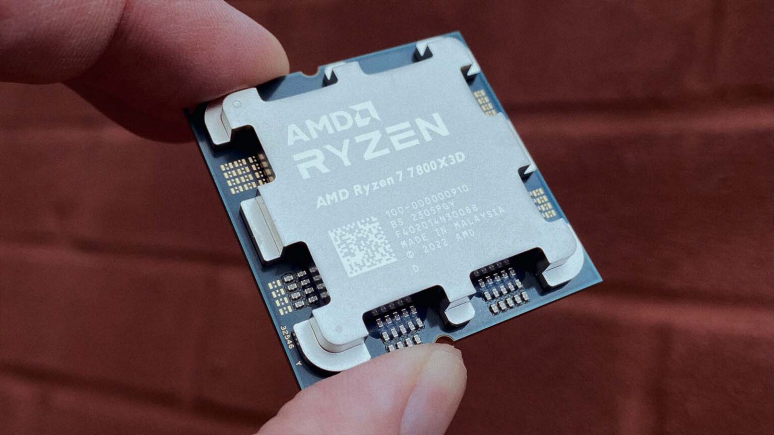 Mão segurando um processador AMD Ryzen 7 7800X3D com o encapsulamento visível, sobre fundo de tijolos avermelhados