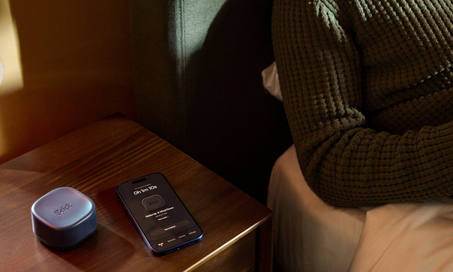 Dispositivo Brick Night ao lado de smartphone na mesinha de cabeceira, com tela mostrando a função de bloqueio de apps, e pessoa deitada na cama ao fundo