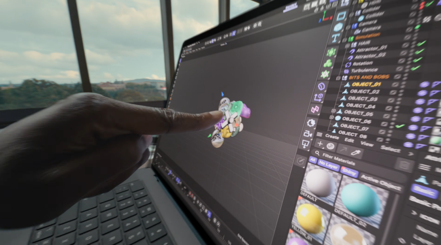 Mão apontando para um agrupamento de esferas coloridas na tela de um notebook com software de modelagem 3D, com lista de objetos e materiais na lateral