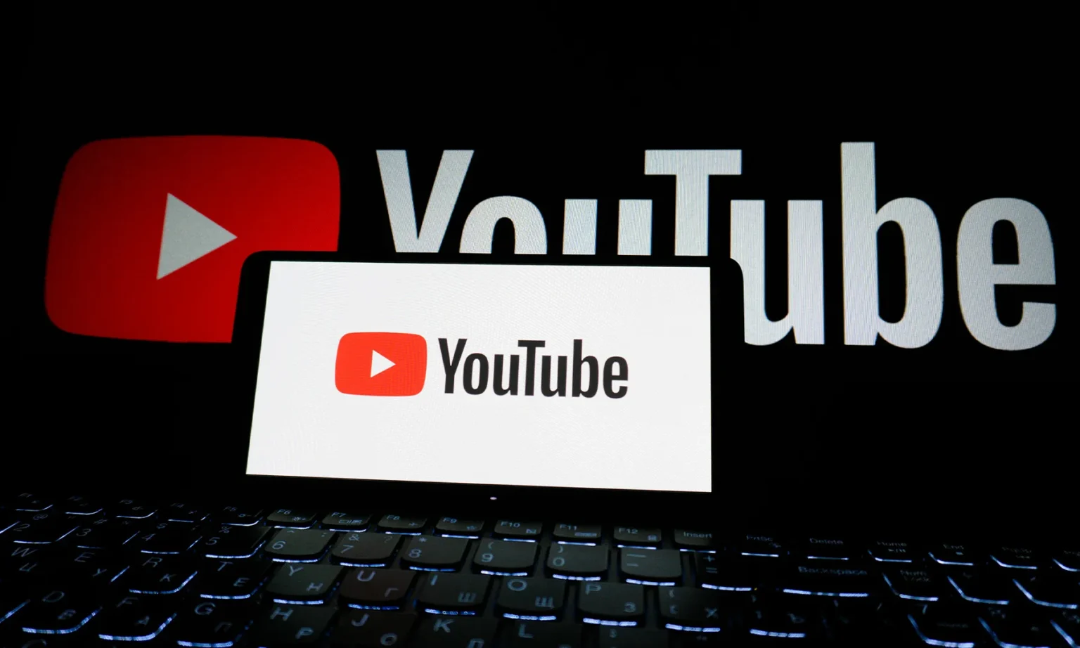 Smartphone com o logo do YouTube sobre um teclado de notebook iluminado, com o logo grande do YouTube ao fundo