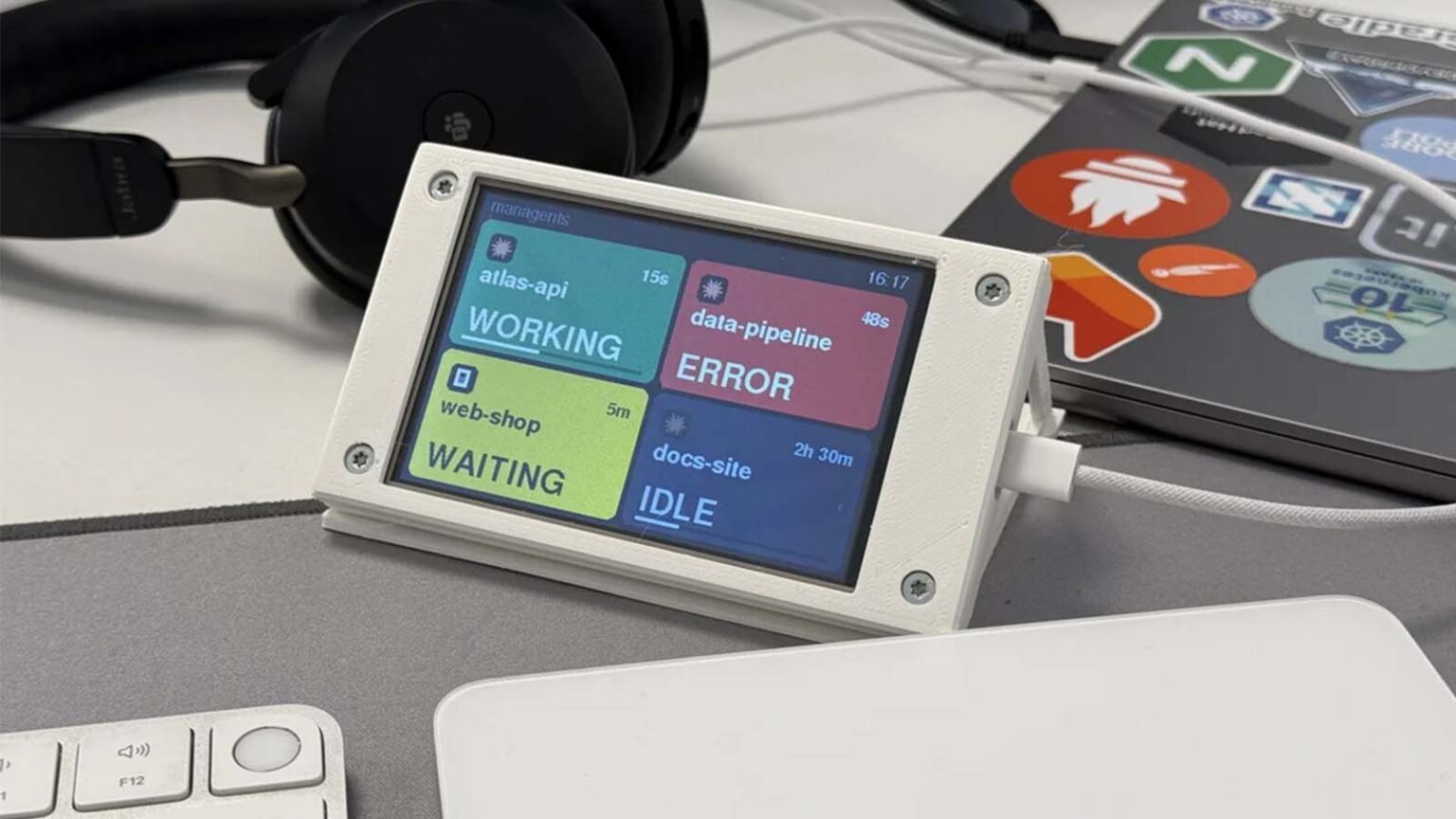Mini display com caixas coloridas mostrando status de agentes de IA (atlas-api WORKING, data-pipeline ERROR, web-shop WAITING, docs-site IDLE) sobre a mesa de…