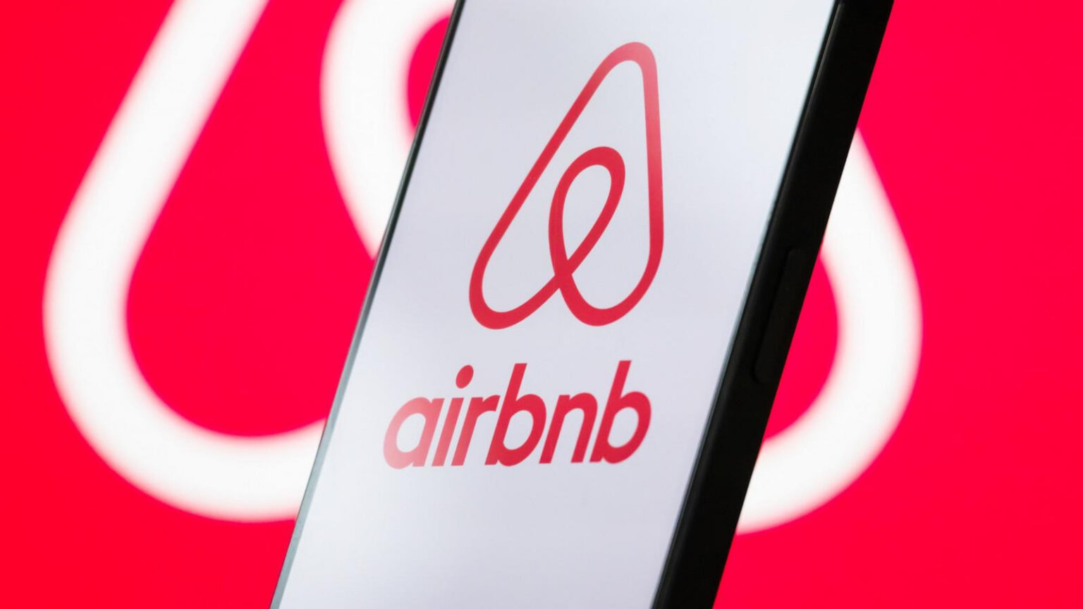 Celular exibindo o logotipo da Airbnb em tela branca sobre fundo vermelho desfocado
