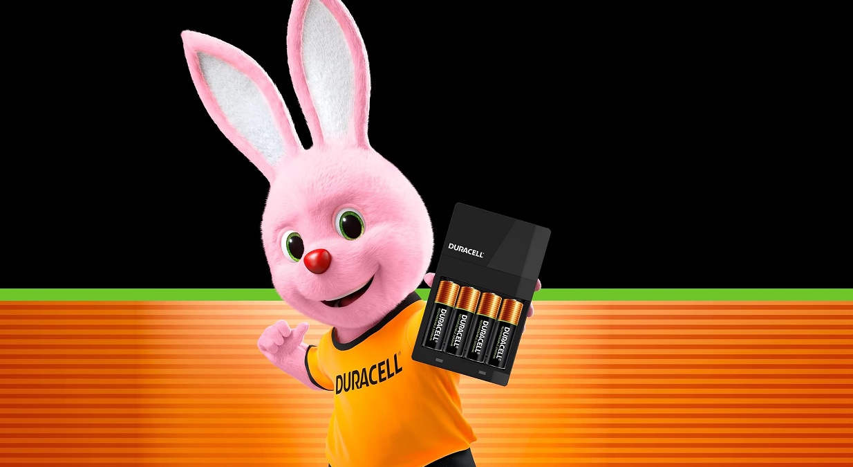 Coelho Duracell de camiseta laranja segurando carregador preto com quatro pilhas AA Duracell encaixadas