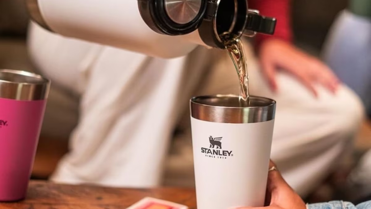 Copo térmico branco Stanley sendo preenchido com líquido por uma garrafa térmica, ao lado de um copo rosa da mesma marca
