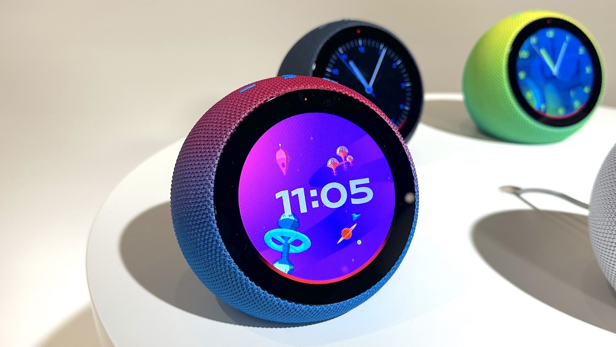 Amazon Echo Time, alto-falante inteligente redondo com tela circular exibindo relógio digital às 11:05, em tons roxos e rosas, ao lado de outros modelos colori…