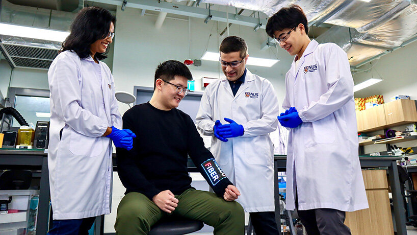 Três pesquisadores de jaleco observam um homem sentado vestindo manga com tecido eletrônico touchscreen, em laboratório da Universidade Nacional de Singapura