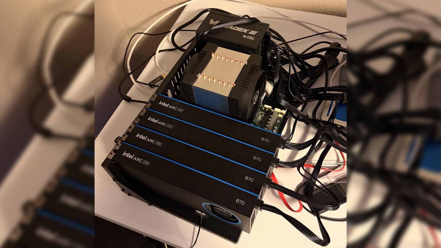 Quatro placas de vídeo Intel Arc B70 empilhadas e interligadas por cabos, com um cooler de torre Thermalright Macho Phantom em cima, montadas sobre uma mesa br…