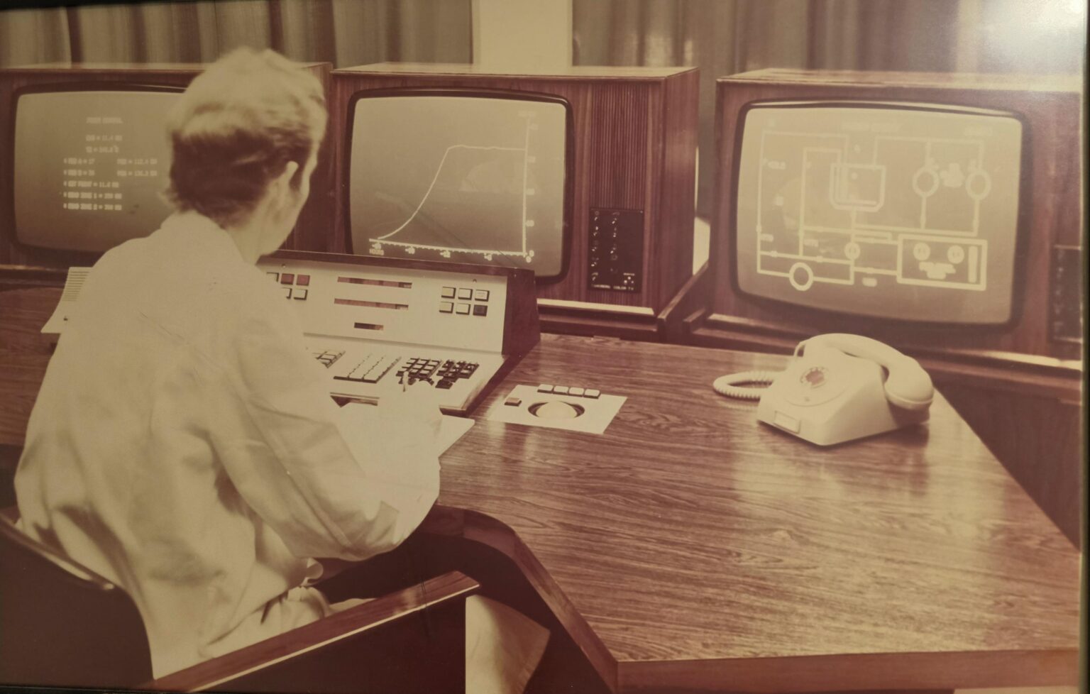 Pessoa de costas operando estação de trabalho com três monitores de tubo de madeira, teclado, painel de controle e telefone de disco, em ambiente dos anos 70